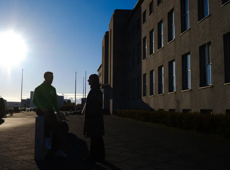 Útlínur af tveimur einstaklingum fyrir framan Aðalbyggingu HÍ, sólin skín