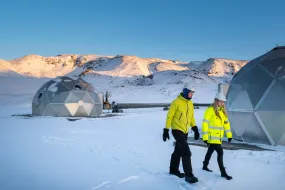 Tvær manneskjur í öryggisbúnaði ganga um snævi þakið rannsóknarsvæði með rannsóknarmannvirkjum og fjöllum í baksýn.
